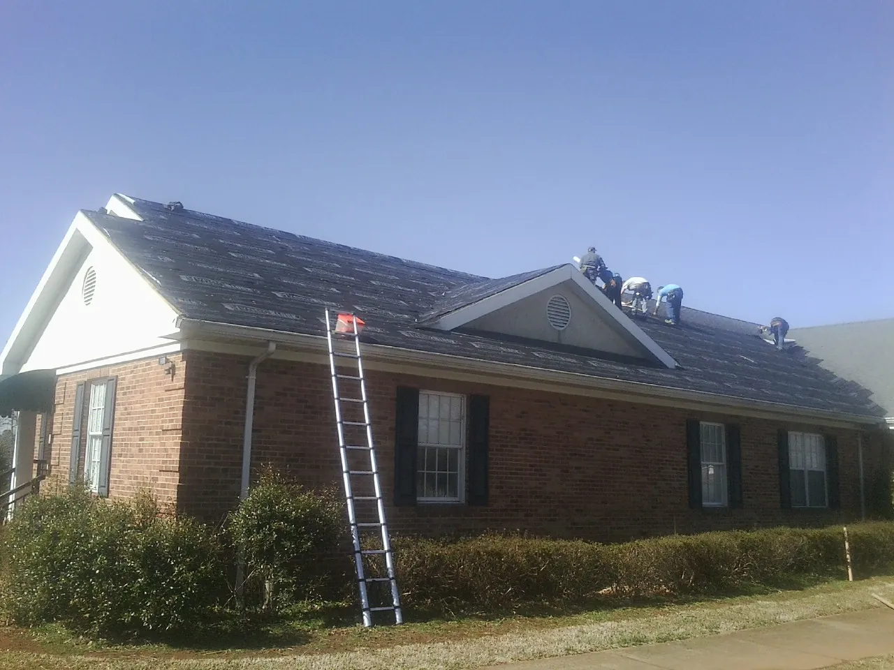 Crew tearing shingles off a brick ranch roof with a ladder set — Emergency Roof Repair work by our crew in Mount Pleasant Mills