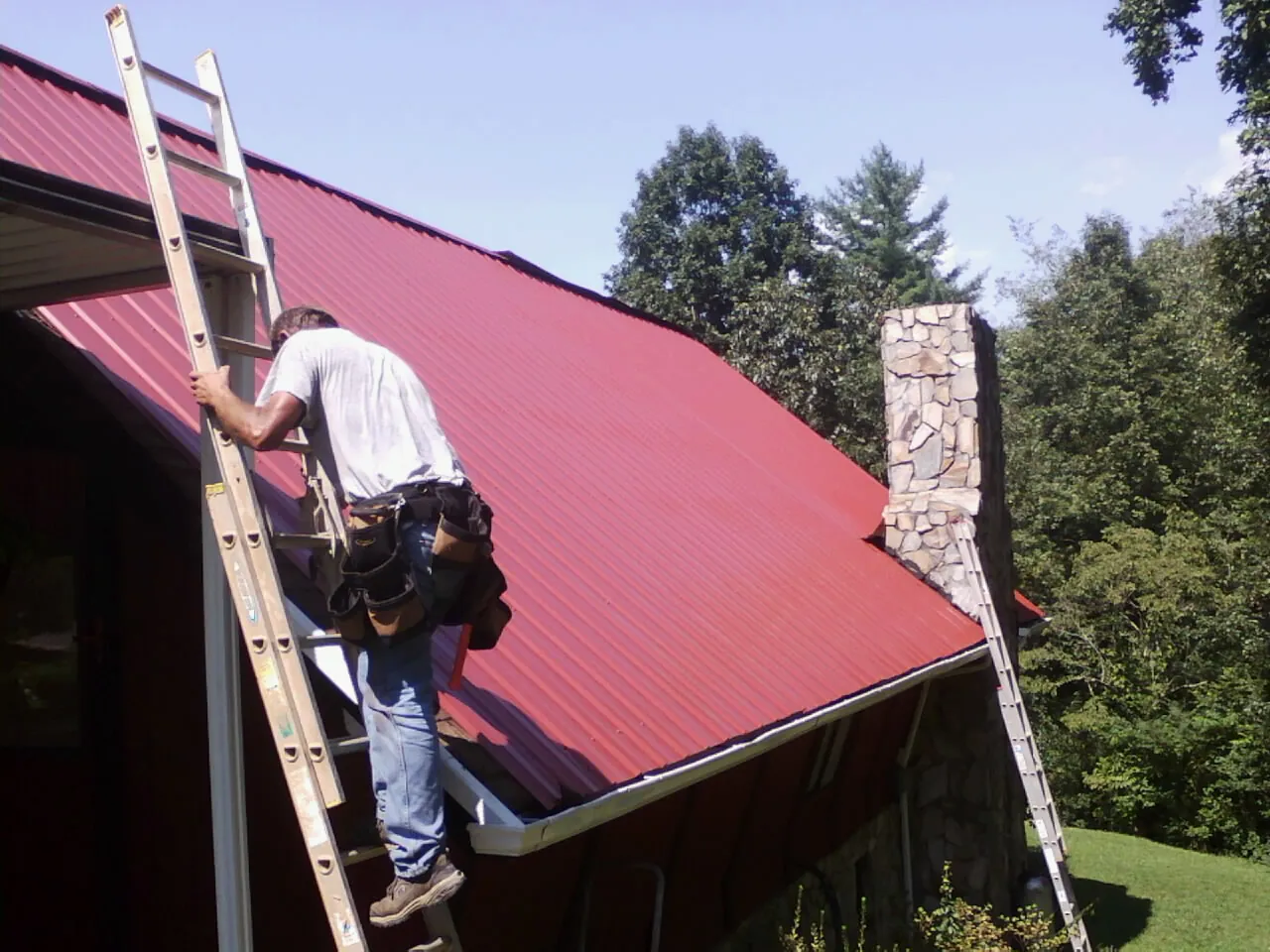 Roofer on a ladder beside a red metal roof and stone chimney — Soffit & Fascia Repair by Roofing Brothers in Mount Pleasant Mills